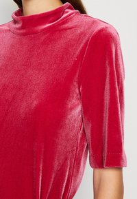 Haut en velours rouge avec un col haut, des manches courtes et un léger éclat ; présente une coupe ample et des coutures subtiles sur les bords des manches.