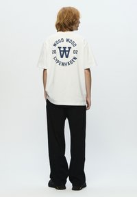 White short-sleeve t-shirt with navy graphic featuring "WOOD WOOD COPENHAGEN" and "2002" on the back. Paired with black wide-leg trousers.