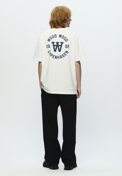 T-shirt bianco a maniche corte con grafica blu navy che presenta la scritta "WOOD WOOD COPENHAGEN" e "2002" sul retro. Abbinato a pantaloni neri a gamba larga.