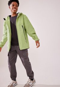 Hellgrüne wasserdichte Jacke mit Kapuze, schwarzen Reißverschlüssen und schwarzem Innenfutter, kombiniert mit grauen Cargo-Hosen mit Taschen und elastischen Bündchen.