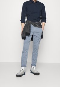 Navy button-up shirt, light blue chinos, and gray-blue sneakers with white laces. Black sweater tied at waist. Simple, streamlined design.