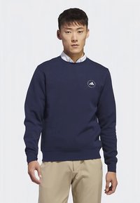 Felpa blu navy in tessuto morbido, con scollo rotondo e polsini a coste. Piccolo logo sul petto. Abbinata a pantaloni beige.
