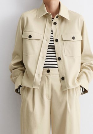 Persona con chaqueta beige abotonada y pantalones beige de cintura alta sobre una camisa de rayas blancas y negras, con las manos en los bolsillos.