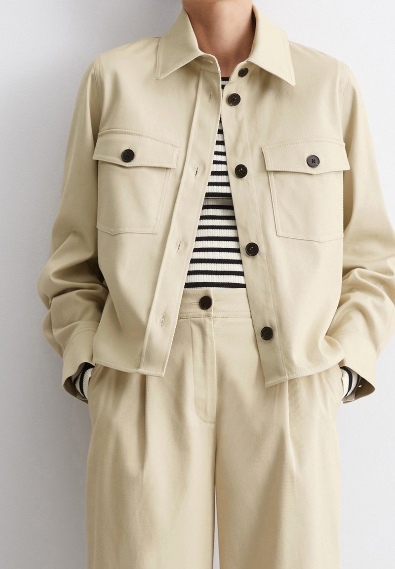 Personne portant une veste beige boutonnée et un pantalon beige taille haute sur une chemise noire et blanche à rayures, mains dans les poches.