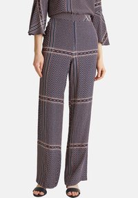 Kvinna klädd i hög midja, vida byxor med geometriska mönster i marinblått, rött och vitt samt svarta sandaler med öppen tå, stående mot en vit bakgrund.