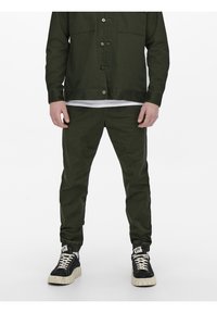 Legergroen button-up shirt en bijpassende broek van gladde stof, gecombineerd met zwarte hoge sneakers met crèmekleurige zolen.