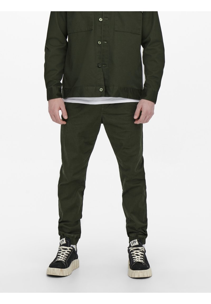 Legergroen button-up shirt en bijpassende broek van gladde stof, gecombineerd met zwarte hoge sneakers met crèmekleurige zolen.