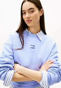 Felpa azzurra con polsini a costine, caratterizzata da un piccolo logo blu "TOMMY JEANS". Sovrapposta a una camicia con colletto a righe bianche e blu.