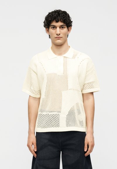 Jeune homme aux cheveux bouclés portant un polo en maille crème à manches courtes et un pantalon foncé, debout devant un fond uni.