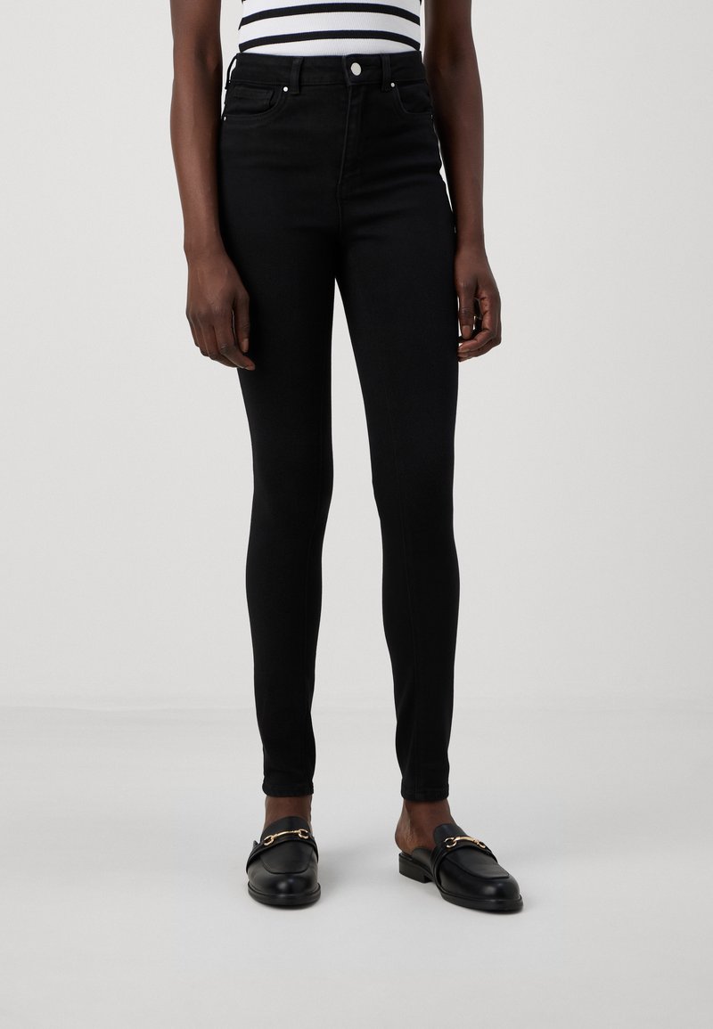Crne skinny traperice visokog struka s glatkom teksturom, pet džepova i metalnim detaljima. Uparene s crnim loafers cipelama sa zlatnim akcentima.