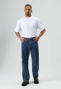 T-shirt blanc col rond, coupe décontractée. Jean en denim bleu avec coutures contrastantes, coupe droite. Baskets noires texturées. Fond neutre.