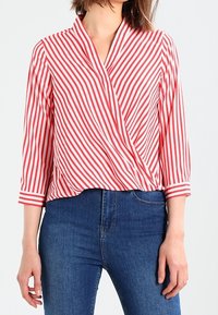 Rød og hvid stribet wrap-bluse med 3/4 ærmer, letvægtsstof, v-hals og løst fit. Båret med blå jeans.