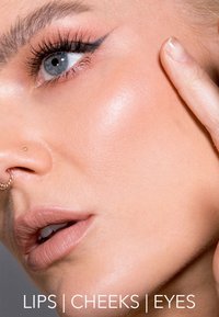 Gros plan sur un visage avec des yeux bleus, un eyeliner bien défini, une peau lisse et des lèvres rose clair, avec un piercings à la cloison nasale et un geste naturel des mains.