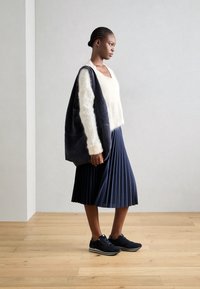 Kvinna som bär en vit fluffig tröja, marinblå veckad kjol, svarta sneakers och bär en stor svart tygväska står på trägolv.