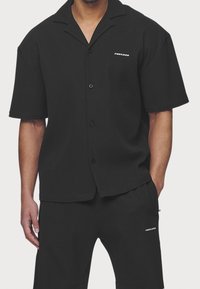 Czarna koszula na krótki rękaw, zapinana na guziki, o prążkowanej fakturze, z płaskim kołnierzem i subtelnym logo na lewej piersi. Zestawiona z pasującymi szortami.