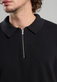 Black ribbed polo shirt featuring a zipper at the collar, with a textured fabric and a structured design.