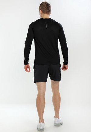 Mann steht mit dem Rücken zur Kamera, trägt ein schwarzes Langarmshirt, schwarze Shorts und weiße Sportschuhe vor weißem Hintergrund.