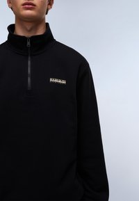 Black zip-up sweatshirt featuring a high collar, textured fabric, and a small logo in gold on the left side of the chest.