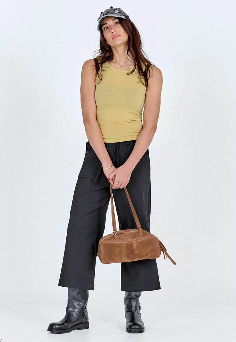 Femme portant un débardeur vert, un pantalon noir à jambes larges, des bottes noires, une casquette grise, tenant un sac à main marron avec les deux mains devant elle.