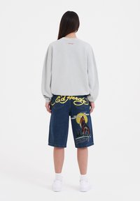 Vaaleanharmaa ylisuurikokoinen collegepaita, jonka takana on huomaamaton logo. Siniset farkkushortsit, joissa on värikäs susigrafiikka ja keltainen kirjonta.