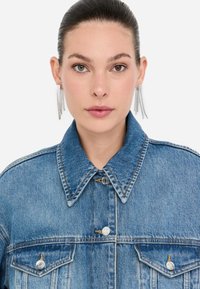 Giacca di jeans in blu chiaro, con colletto classico, due tasche sul petto e bottoni argentati. Il modello indossa orecchini a catena lunga argentati.