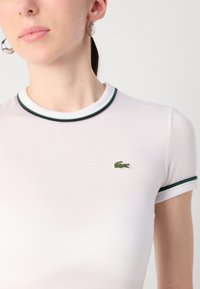 Weißes, tailliertes T-Shirt mit grünem und marineblauem geripptem Kragen und Ärmelabschlüssen. Kleines grünes Krokodil-Logo auf der Brust gestickt.