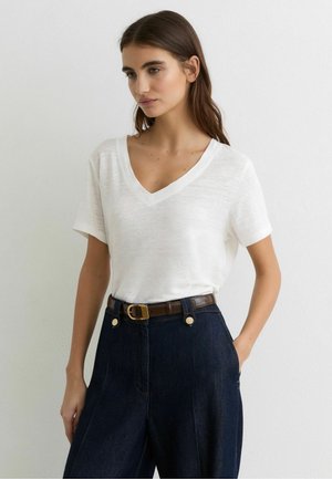 Fiatal nő fehér V-nyakú rövid ujjú ingben, melyet magas derekú sötétkék farmerral és barna övvel visel, egy sima fal előtt áll.