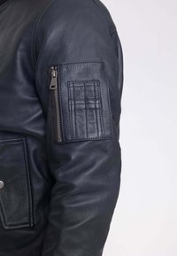 Donkerblauwe leren jas met een ritszak op de linker mouw en een opgestikte zak aan de voorkant. Glad textuur en klassiek ontwerp.