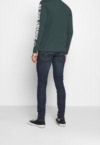 Persona con camisa de manga larga verde oscuro con "SÉ EL CAMBIO" en la manga, jeans azul oscuro y zapatillas Converse negras, de pie de espaldas.