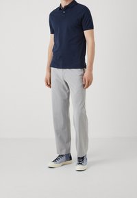 Mann in navyblauem Poloshirt, hellgrauen Hosen und blauen High-Top-Sneakern, der vor einem schlichten weißen Hintergrund steht.