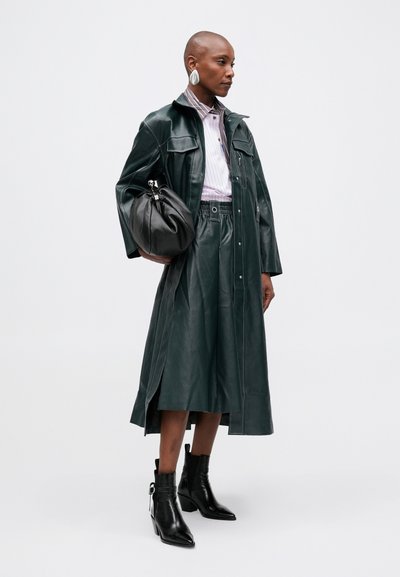 Casaco longo de pele verde com grandes bolsos, combinado com uma camisa às riscas e uma saia de cintura alta. Botas pretas de tornozelo com salto robusto.