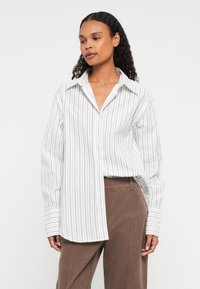 Vrouw met lang krullend haar, gekleed in een witte blouse met lange mouwen met zwarte verticale strepen en een bruine corduroy broek, staand tegen een witte achtergrond.
