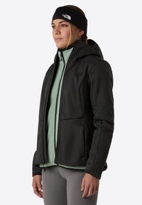 Svart jacka med huva, dragkedjestängning, med en strukturerad yta. Bärs över ett ljusgrönt lager och parats med grå leggings.