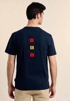 Camiseta de algodón azul marino con un gran número "3" bordado en bloques delineados de rojo, amarillo y verde en la parte posterior. Manga corta.