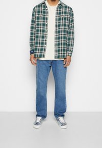 Rutigt skjorta i grönt och kräm över en ribbad vit t-shirt. Blå jeans och mönstrade sneakers fulländar outfiten. Avslappnad passform och lager-på-layer-look.