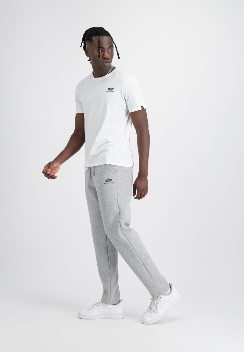 Homme portant un t-shirt blanc et un pantalon de jogging gris avec des baskets blanches, debout et regardant vers sa gauche sur un fond clair uni.