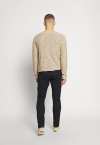 Muž stojící zády, oblečený v béžovém svetru, černých džínách a béžových teniskách, na jednobarevném bílém pozadí.