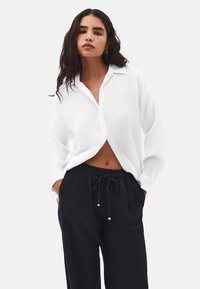 Camicia bianca con colletto, indossata aperta sopra pantaloni neri con cordino. La camicia ha una vestibilità comoda e un tessuto leggero.