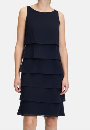 Femme portant une robe bleu marine sans manches avec cinq volants horizontaux superposés du haut jusqu’au genou.