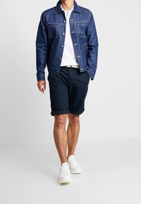 Jeansjacka med vit sömnad, mörkblå shorts och vita sneakers. Jackan har knäppt stängning och en klassisk krage.