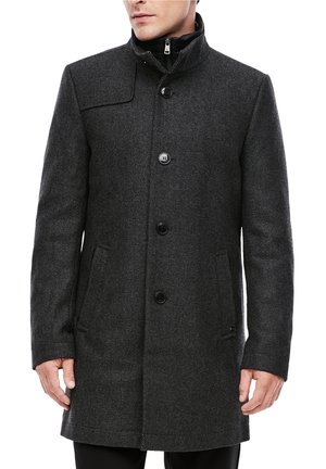 Man wearing a dark gray tailored wool coat with front buttons, side pockets, and a high collar over a black zip-up inner layer.