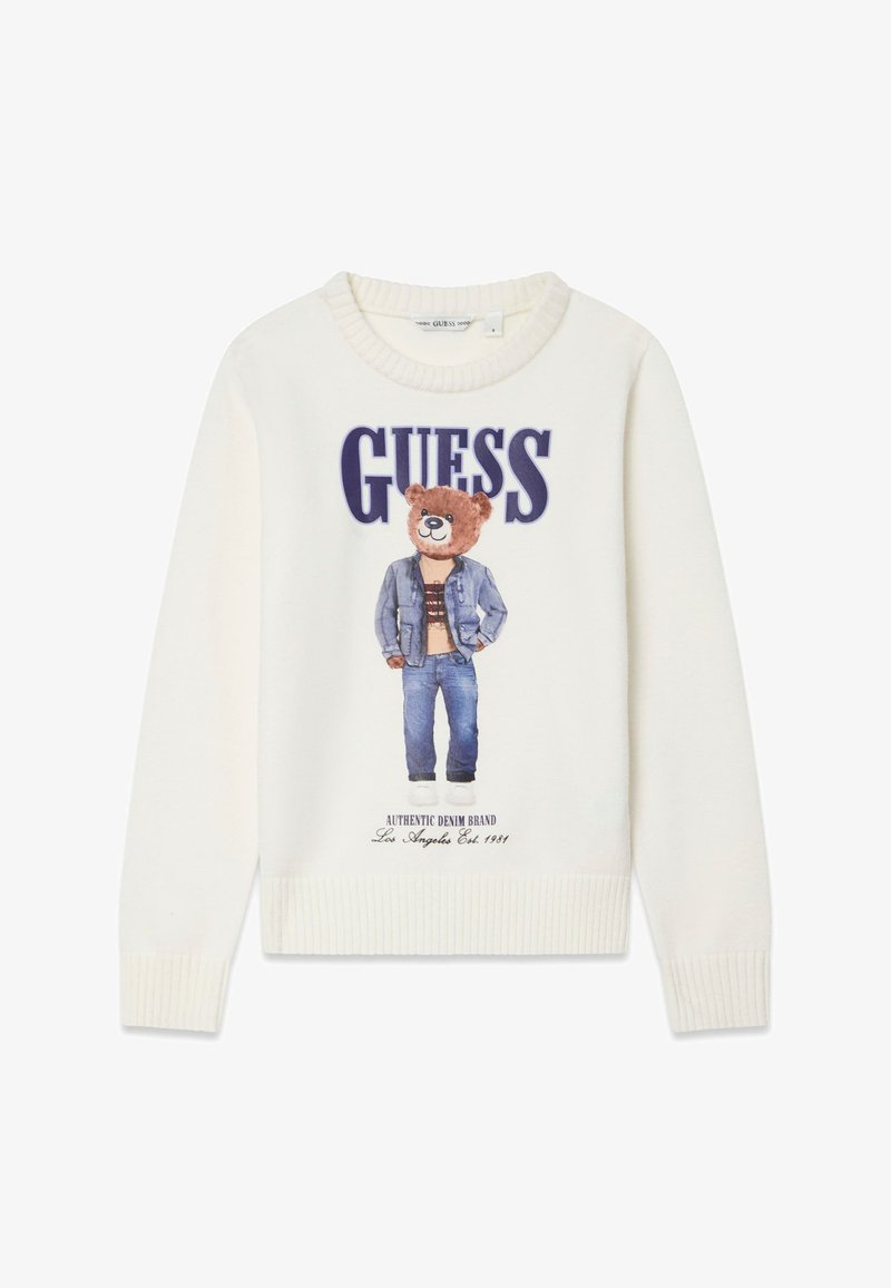 Pull blanc avec un imprimé d'ours portant une veste en jean et un jean, avec le texte « GUESS » au-dessus et des poignets et un ourlet côtelés.