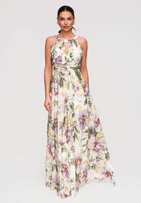Femme portant une robe longue sans manches à col licou avec des motifs floraux verts, violets et jaunes sur fond blanc.