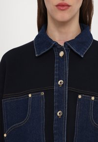 Camicia in denim e maglia con colletto e tasche in denim blu scuro, corpo nero, bottoni dorati e dettagli di cucitura a contrasto.