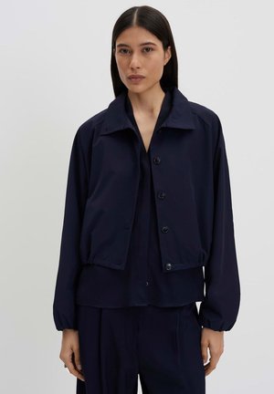 Frau mit langen dunklen Haaren trägt eine dunkelmarineblaue, geknöpfte Jacke über einem passenden Hemd und Hose, stehend vor einem einfarbigen hellen Hintergrund.