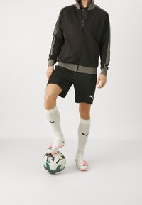 Giacca da ginnastica nera con accenti grigi, pantaloni corti neri, calzini bianchi e scarpini da calcio colorati, in piedi su un pallone da calcio.