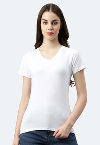 Junge Frau mit langen braunen Haaren, die ein schlichtes weißes V-Ausschnitt-T-Shirt und Bluejeans trägt, steht vor einem hellen Hintergrund.