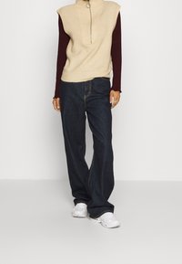 Jersey sin mangas de color beige con canalé y cuello alto, combinado con pantalones vaqueros de pierna ancha de mezclilla oscura. Zapatillas deportivas blancas completan el conjunto.