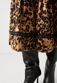 Abito con stampa leopardata su una base marrone dorato e macchie nere, caratterizzato da una fascia nera con occhielli. Abbinato a stivali neri in pelle al ginocchio.