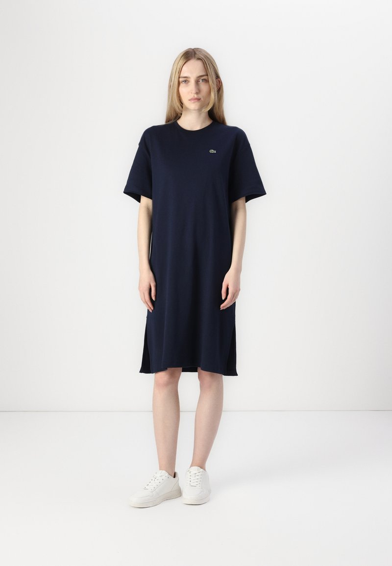 Navy cotton T-shirt dress with short sleeves and side slits, featuring a small green logo on the chest. Paired with white sneakers.
